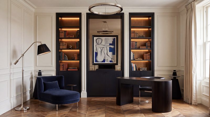 Study shown as designed, with dark lit shelving, a sculptural desk and a navy chair. A visualisation.
