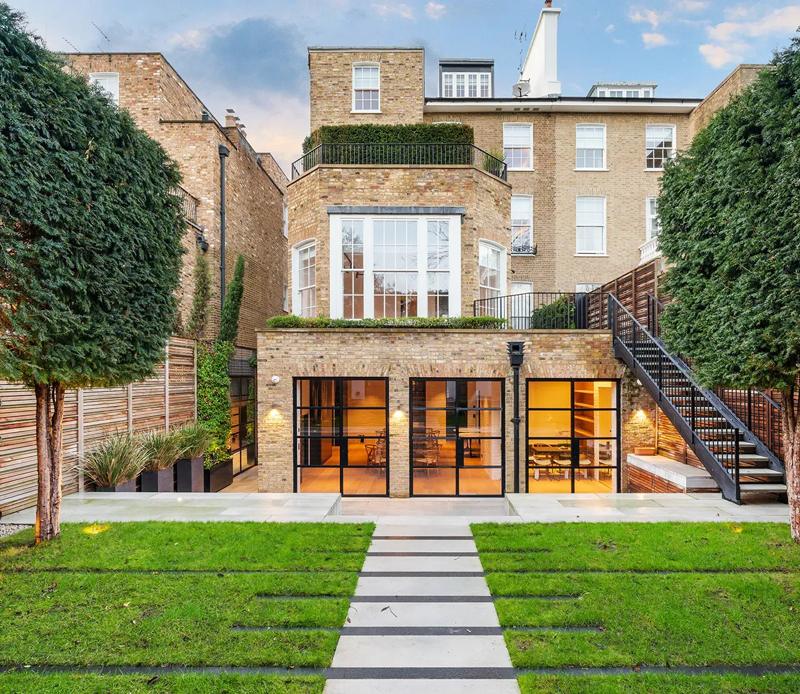 The garden front of a Chelsea house built by NU Projects: a brick lower-ground extension with black steel doors lit from inside, a planted terrace above, and a stone path across the lawn.