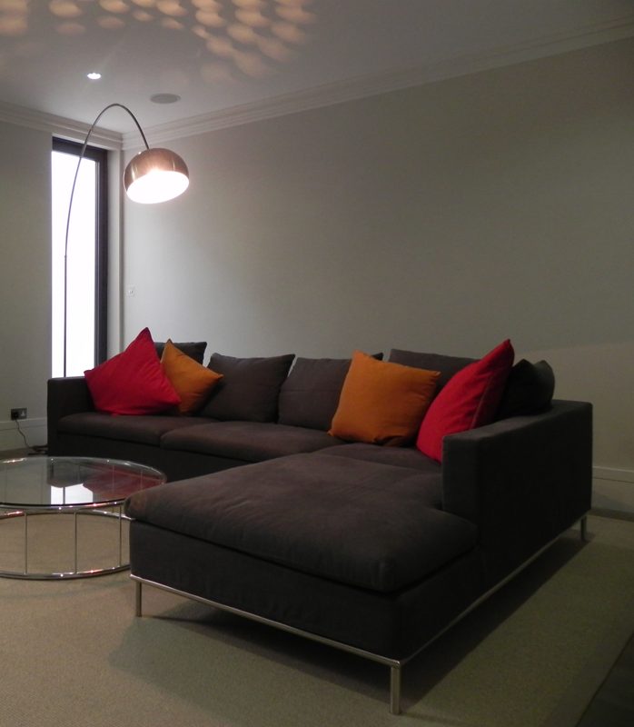 Sitting room with a tall narrow full height window, a grey corner sofa, a round glass table and an arc floor lamp.