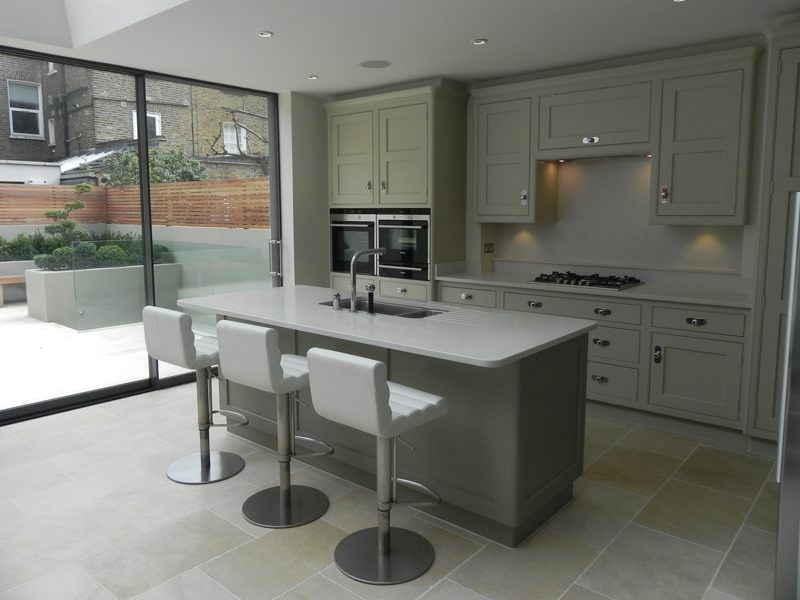 Painted kitchen with a stone topped island and three white bar stools, beside full height sliding glass onto a paved courtyard garden.