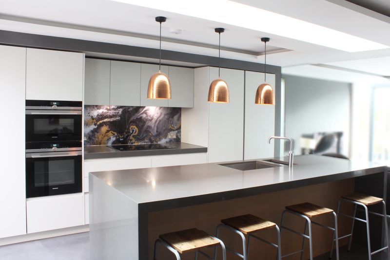 Open plan London kitchen with a flush ceiling raft and a long rooflight slot. Three copper pendants hang over a pale island, with no visible cooling units.