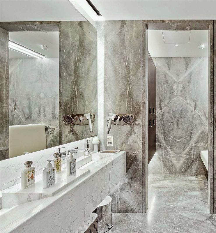 Finished London apartment bathroom in grey book matched stone, with a white stone basin, a linear ceiling grille and a walk in shower beyond.