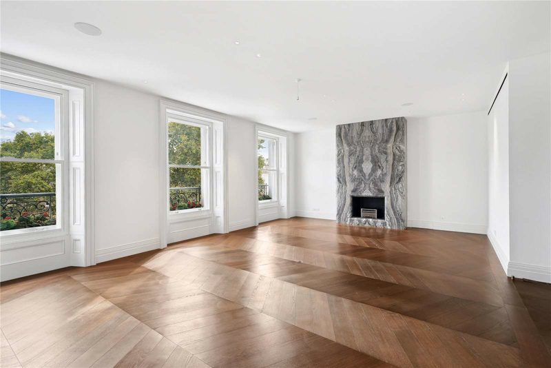 Empty London apartment reception room with tall sash windows onto trees, a chevron oak floor, flush ceiling speakers and a stone fireplace.