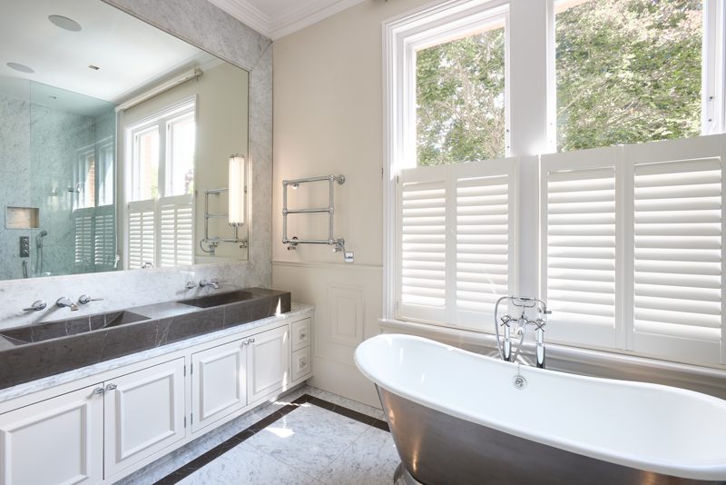 White bathroom with a roll top bath below timber shutters.