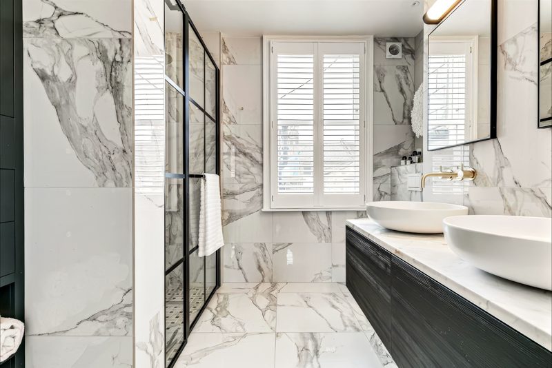 Bathroom in book-matched marble with twin basins and a black-framed shower.