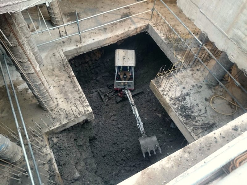 A small digger at the bottom of the excavation, seen from above. Piles and steel surround it.