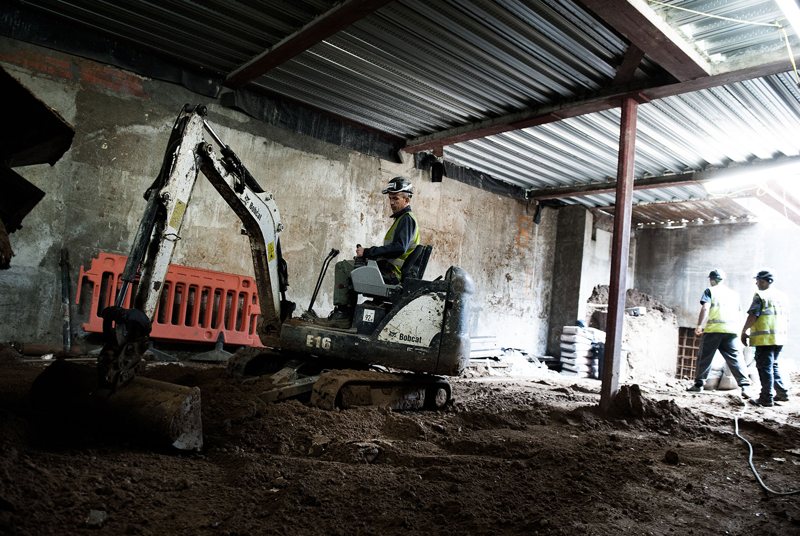 A mini excavator digging out a basement under temporary steel propping.