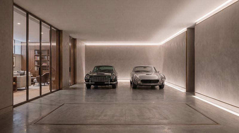 A private car gallery below a London house: two classic cars, a flush car lift in a polished concrete floor, and a glass wall to a walnut lounge.