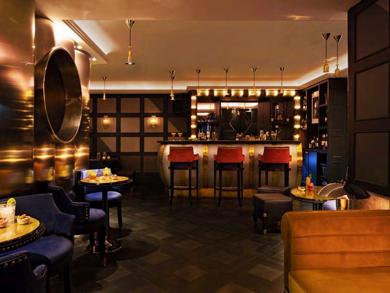 A cocktail bar with red leather stools, a lit back bar of mirrors and bulbs, and navy armchairs.