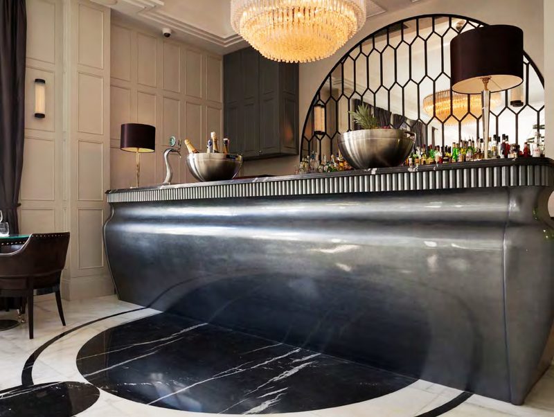 A curved metal-clad hotel bar on a marble floor, with an arched lattice screen and a chandelier behind.