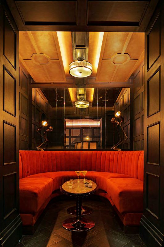 A booth in deep orange velvet under a gold-lit ceiling, with a round brass table between dark panelled walls.
