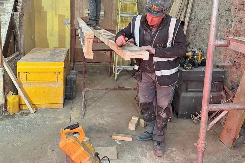 A carpenter marking timber on site, tools and props behind him.