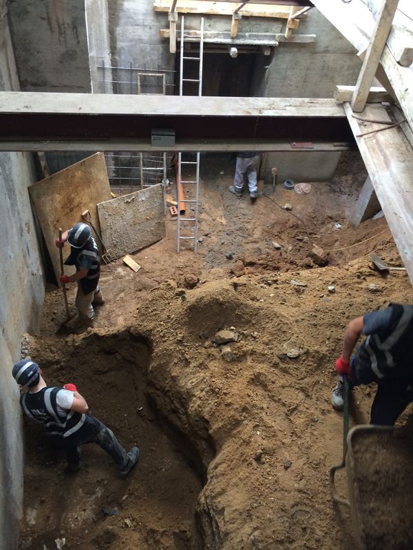 Three of the site team digging out a basement under a steel beam, with temporary props and a ladder behind.